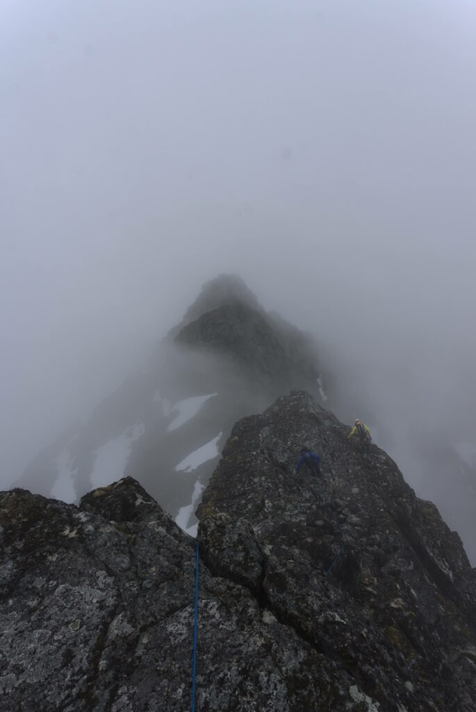 View from a mountain ridge of the way ahead; a rope goes over the edge and there are two climbers below; ahead the ridge appears out of the cloud
