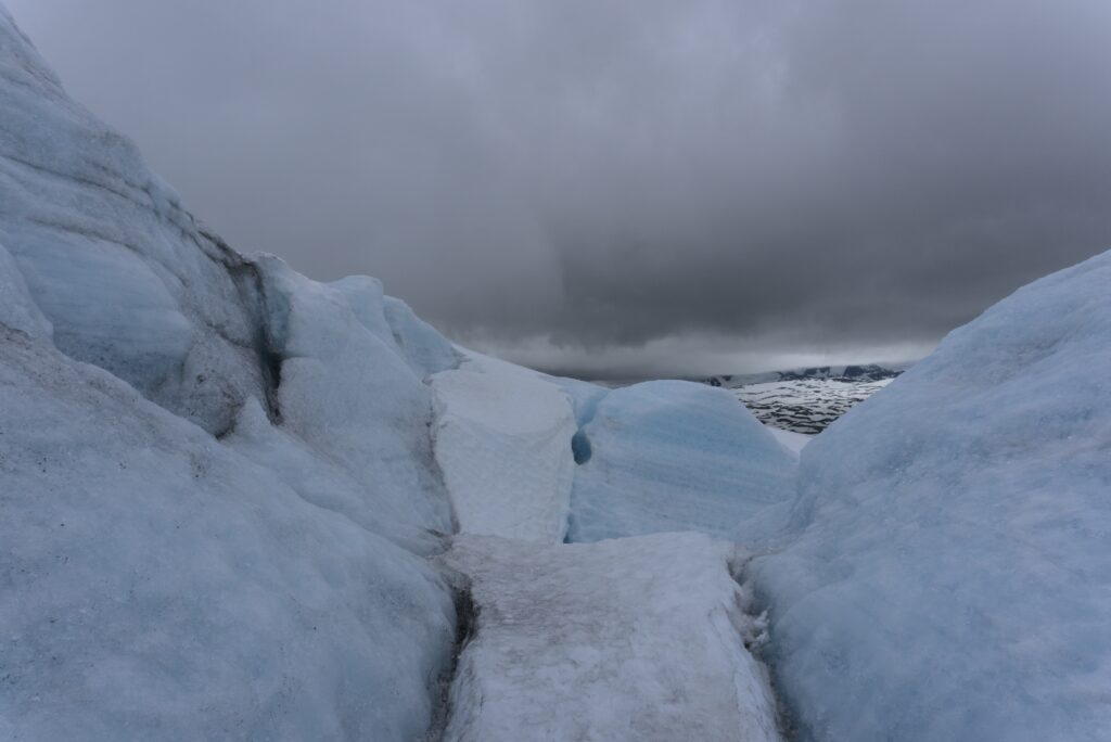 View from inside a large crevasse on a glacier; a landscape of ice with many shades of blue and grey; a stormy sky above