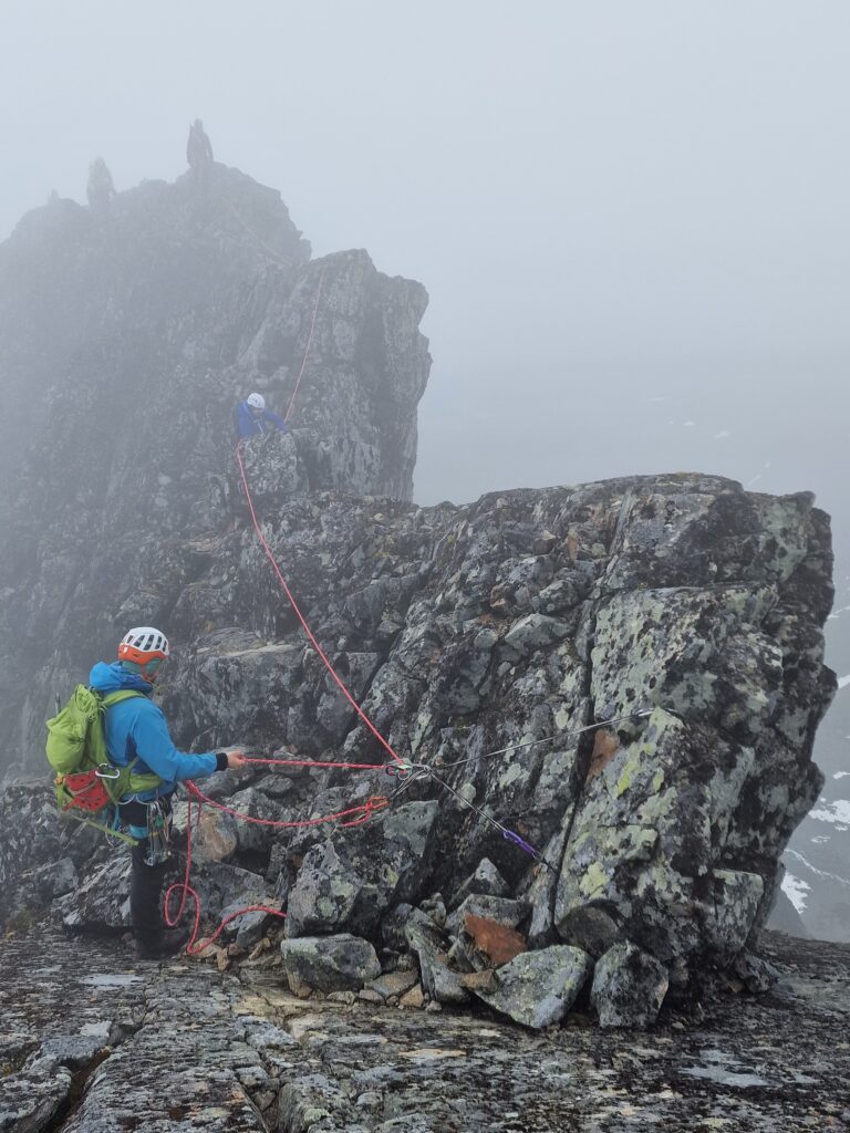 Someone belaying other climbers over from a top anchor on a mountain ridge in cloudy weather
