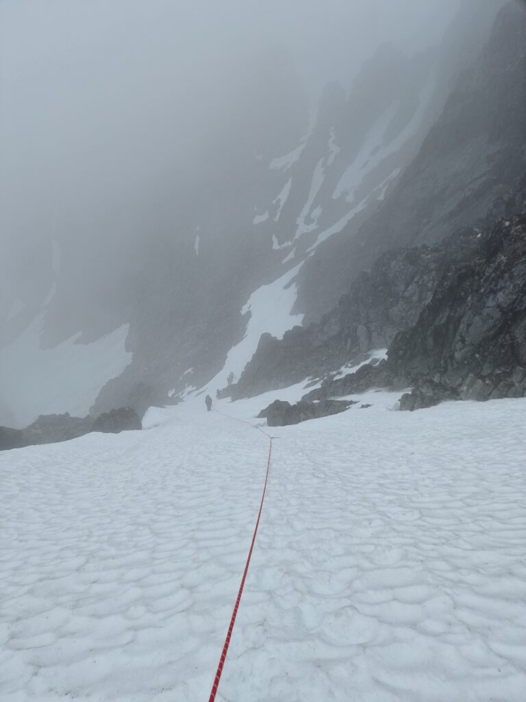 View down a steep snowy slope; climbing down on a rope; people in the clouds ahead