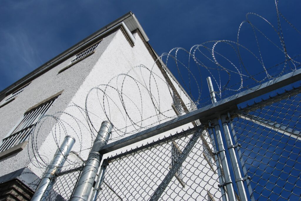 View up from outside a prison, a takk fence with barbed wire, a building, and a blue sky