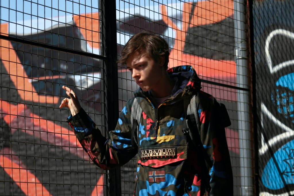 An adolescent boy in a colourful jacked facing the camera and looking downwards in a thoughtful manner. He has his fingers through the holes in a tall metal fence. Colourful background with graffiti on a tall wall.
