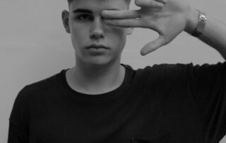 Black and white picture of an adolescent or young man looking straight at the camera. He's wearing a black T-shirt and he's holding his left hand over his left eye in the shape of a gun. He looks serious.