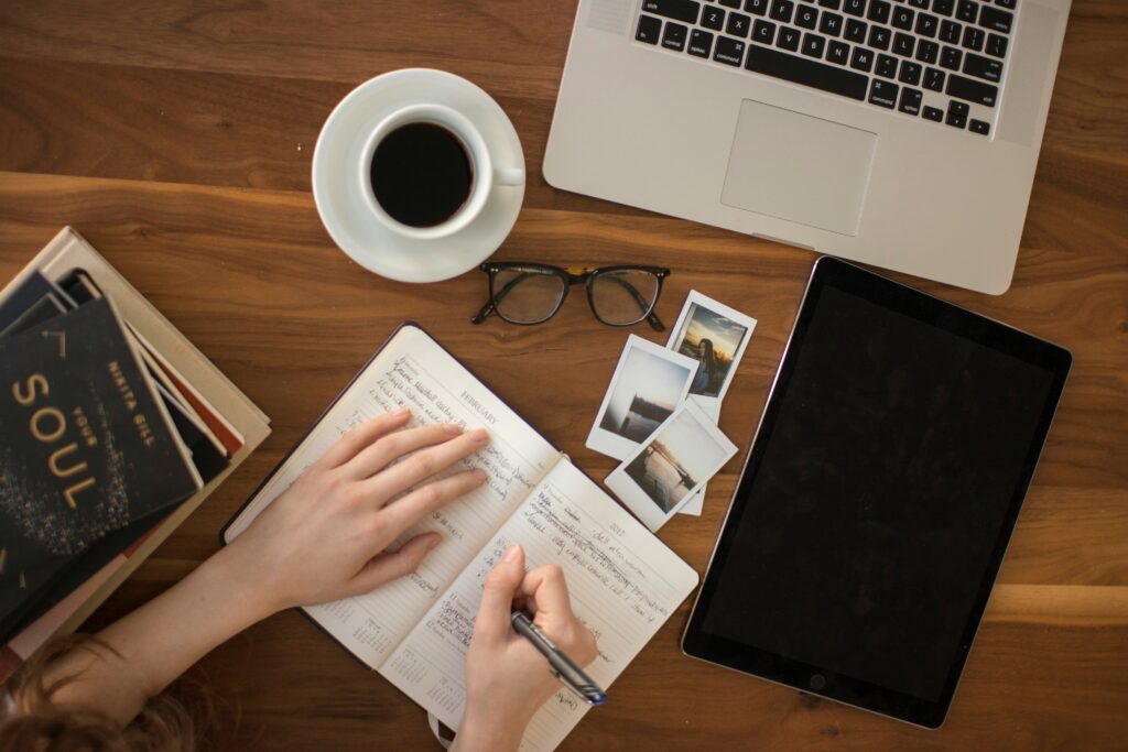 Picture of someone writing in a diary, with an open laptop, some photographs, a cup of coffee and some glasses on a wooden table